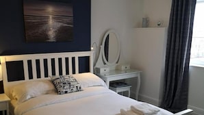 A neatly made bed with white linens and folded towels, a bedside table with a lamp, a dresser with a mirror, and a painting on the wall. - The Sunfold (Weston-super-Mare)