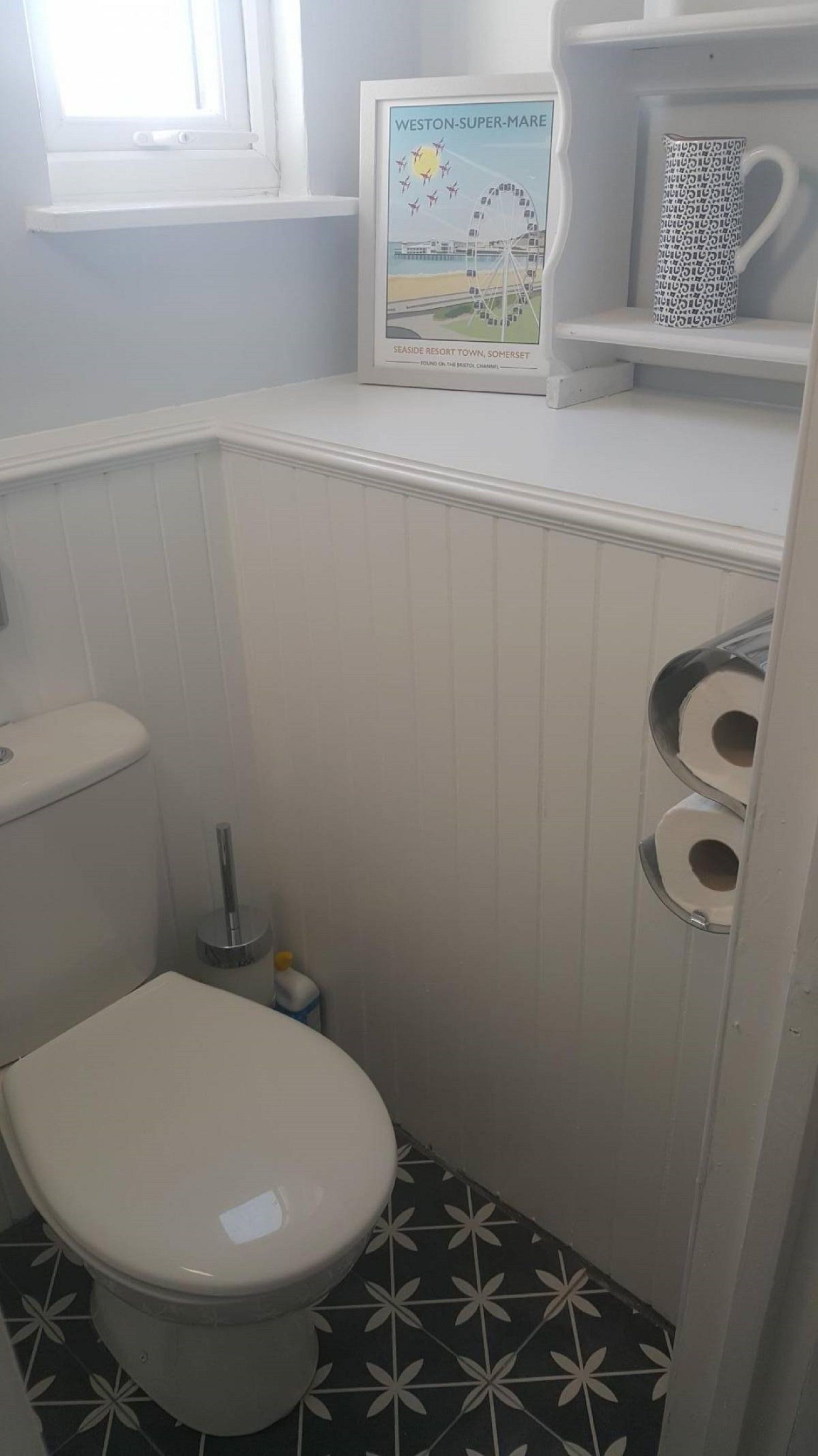 Een kleine, witte badkamer met een toilet, een rol toiletpapier en een lijst met een foto van Weston-Super-Mare aan de muur.