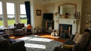 A traditional living room with a fireplace, patterned furniture, and a large window overlooking a lawn. - The Sunfold (Weston-super-Mare)