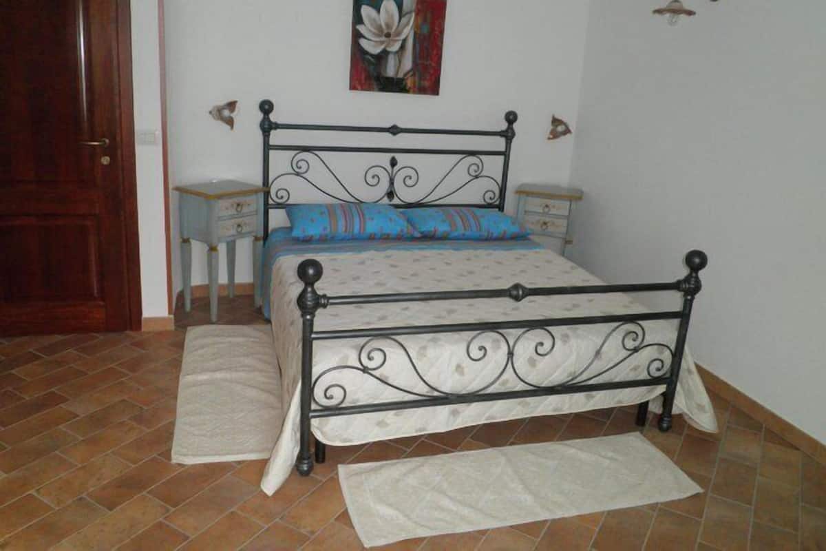 Una camera da letto con una struttura del letto in metallo, un letto singolo con copripiumino decorato, due comodini con lampade, una porta in legno e un pavimento piastrellato.