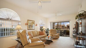 A spacious living room with a large sofa, a glass coffee table, and a comfortable armchair. There is a desk with a computer and a bookshelf filled with books. The room has large windows with a view of the outside.
