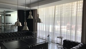 Een moderne woonkamer met een zwarte leren bank, een grote zwarte salontafel en drie hanglampen. Er is een groot raam met dunne gordijnen en uitzicht op buiten.