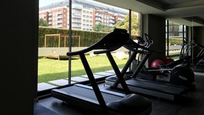 Een sportschool met loopbanden en trainingsapparatuur, grote ramen en uitzicht op een gebouw en groen buiten.