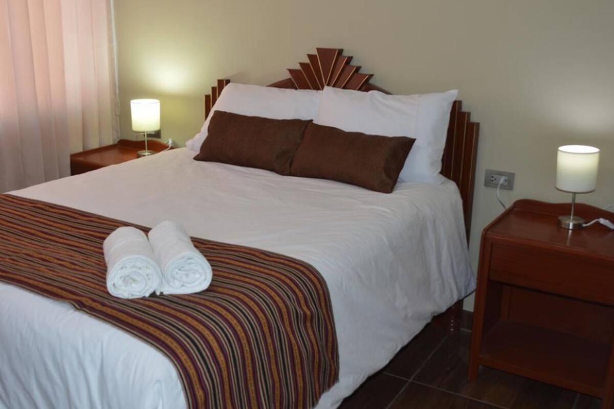 A neatly made bed with a striped bedspread, two rolled towels, and a wooden headboard. There are two bedside lamps and a nightstand with a lamp.