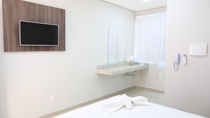 Quarto de hotel com televisão de ecrã plano na parede, casa de banho com lavatório e espelho, e cama com roupa de cama branca.