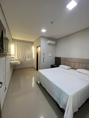 Una habitación de hotel con cama, mesita de noche, televisor montado en la pared y ventana con cortinas.