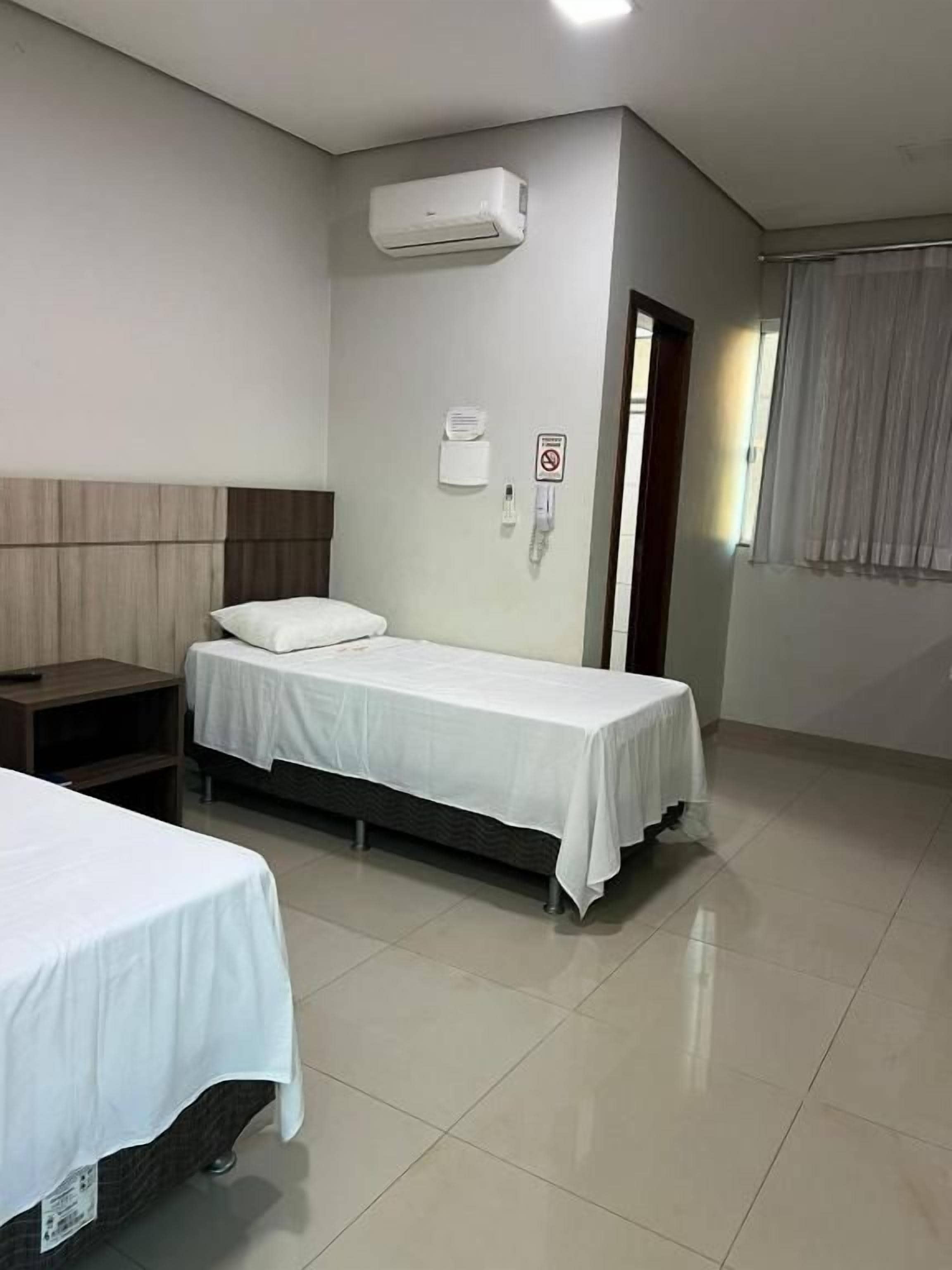 A hotel room with two beds, a wooden headboard, a white air conditioning unit, a bedside table, and a window with curtains.