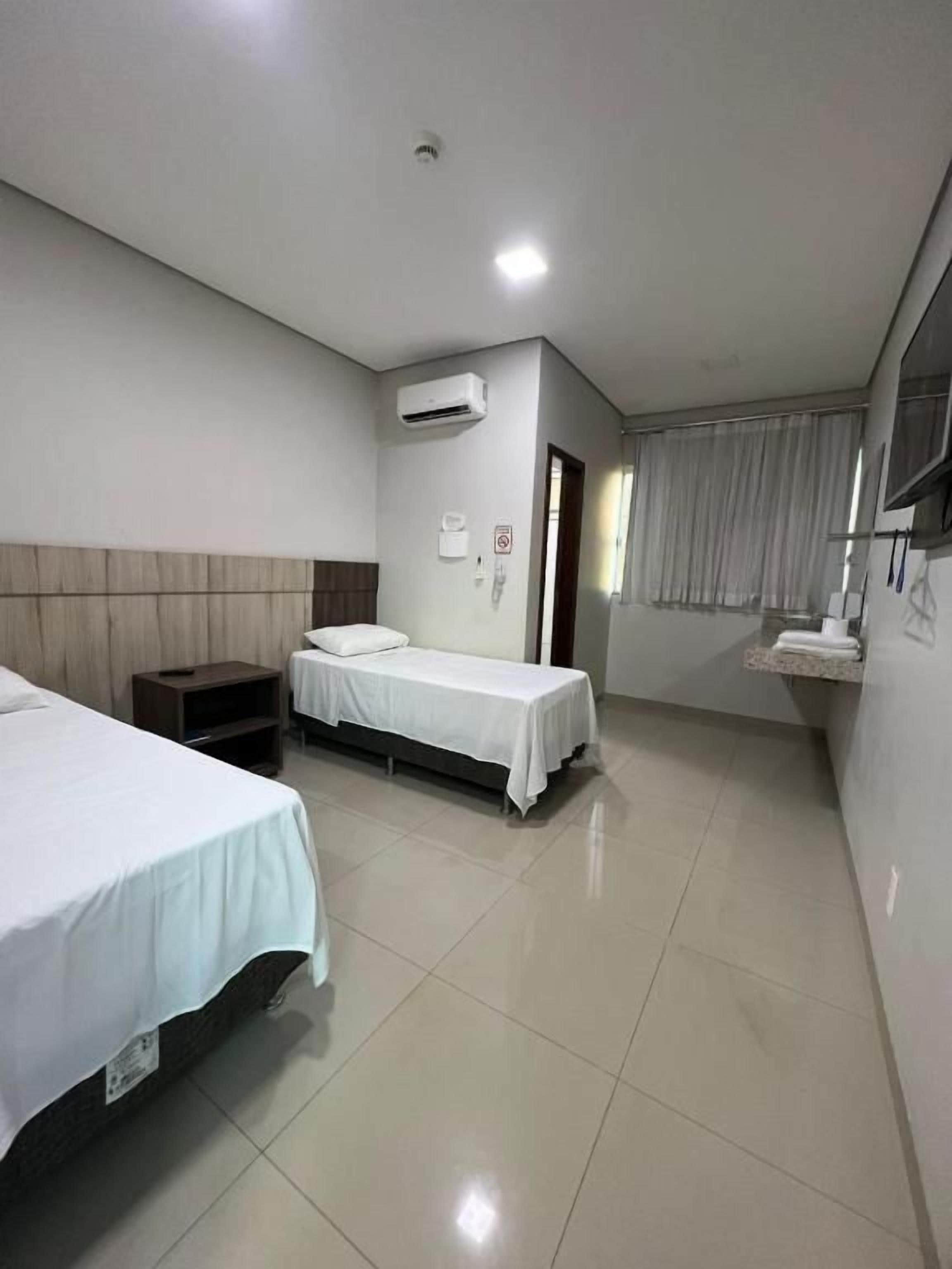 A hotel room with two beds, a bedside table, an air conditioning unit, a window with curtains, and a wall-mounted TV.