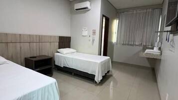 A hotel room with two beds, a bedside table, an air conditioning unit, a window with curtains, and a wall-mounted TV.