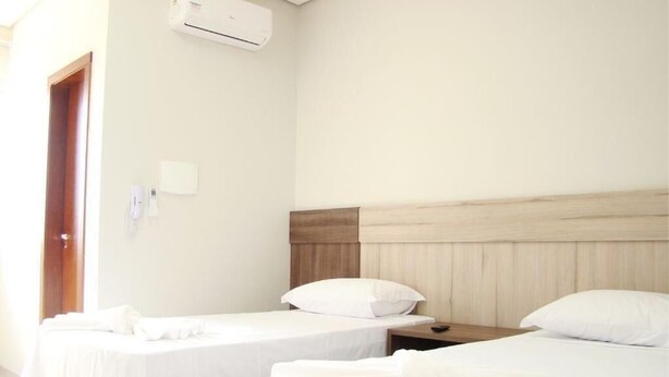Quarto de hotel com duas camas de solteiro, cabeceira de madeira, mesa de cabeceira e ar condicionado.