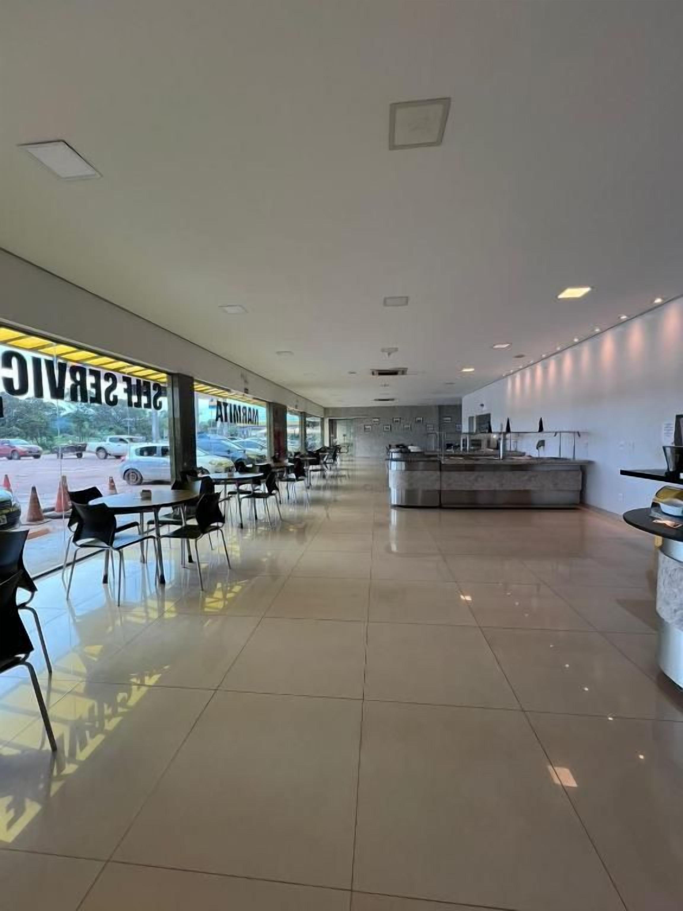 Un amplio espacio interior con una larga fila de mesas y sillas, un mostrador de recepción y vistas al exterior.