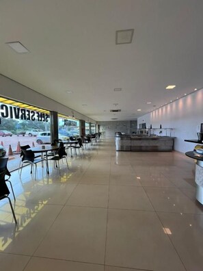 A spacious indoor area with a long row of tables and chairs, a reception desk, and a view of the outside. - Hotel PetroShop (Palmas)
