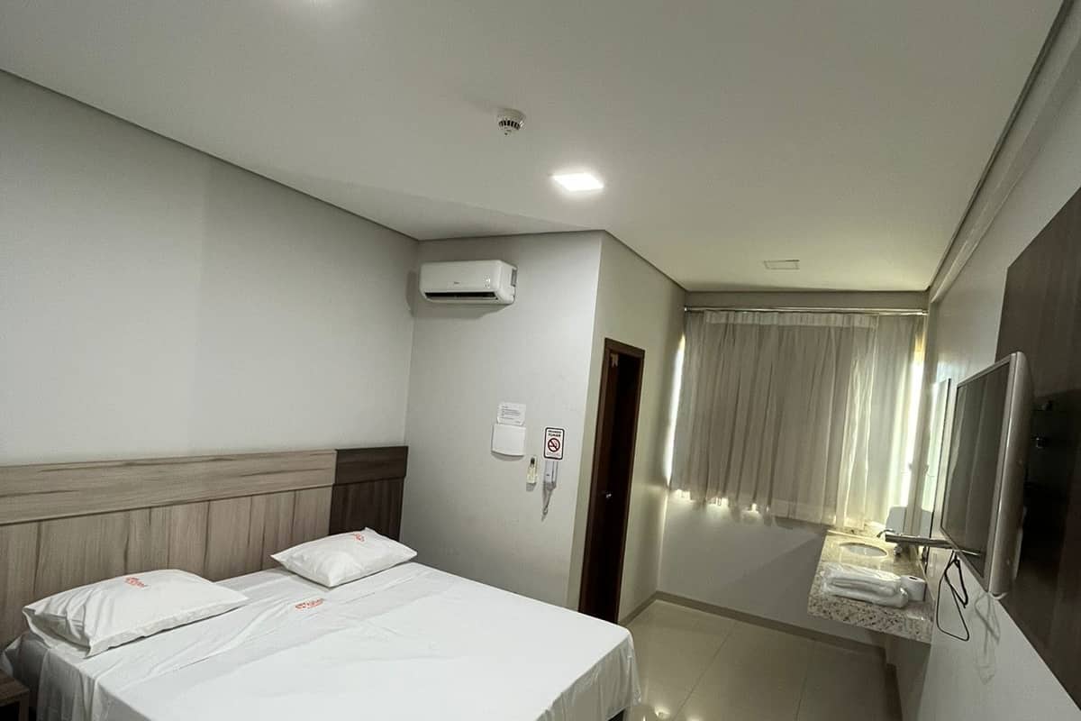 A hotel room with a large bed, a TV, a bathroom with a mirror, and an air conditioning unit.