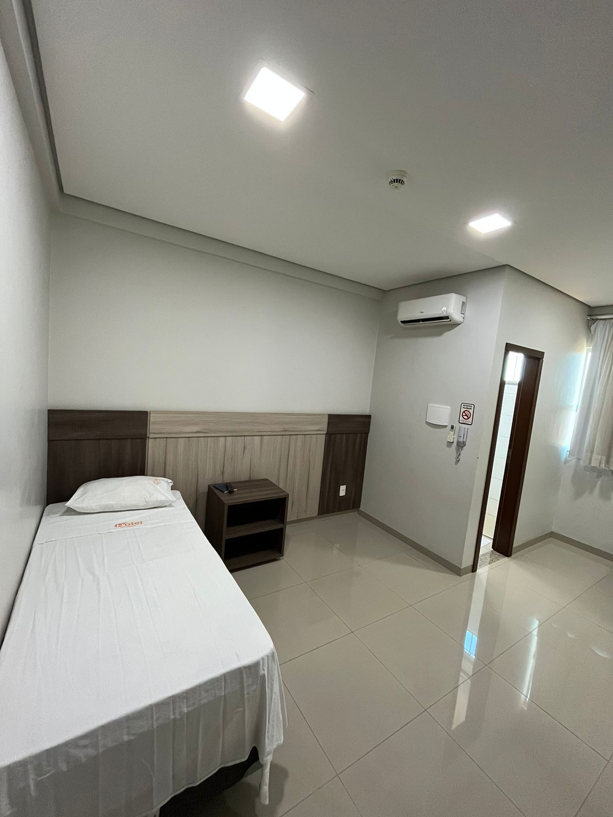 A hotel room with a bed, a nightstand, a wall-mounted air conditioner, a door with a no-smoking sign, and a window with curtains.