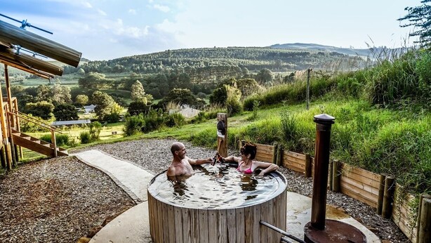 Un jacuzzi con revestimiento de madera, rodeado de una terraza de madera y un sendero de piedra, con vistas a una ladera panorámica.