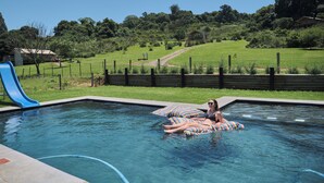 Una piscina con tobogán, rodeada de césped y una colina al fondo.