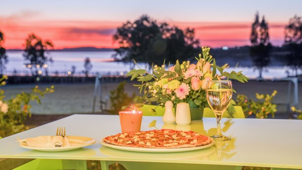 A table set with a pizza, a candle, and a glass of wine, with a view of the sunset over water. - The Anchorage (Vaal Marina)