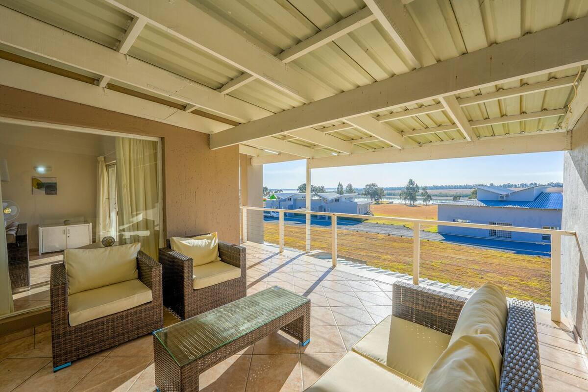 Uma área de estar coberta ao ar livre, com móveis de vime e uma mesa de vidro, com vista para um campo esportivo e edifícios.