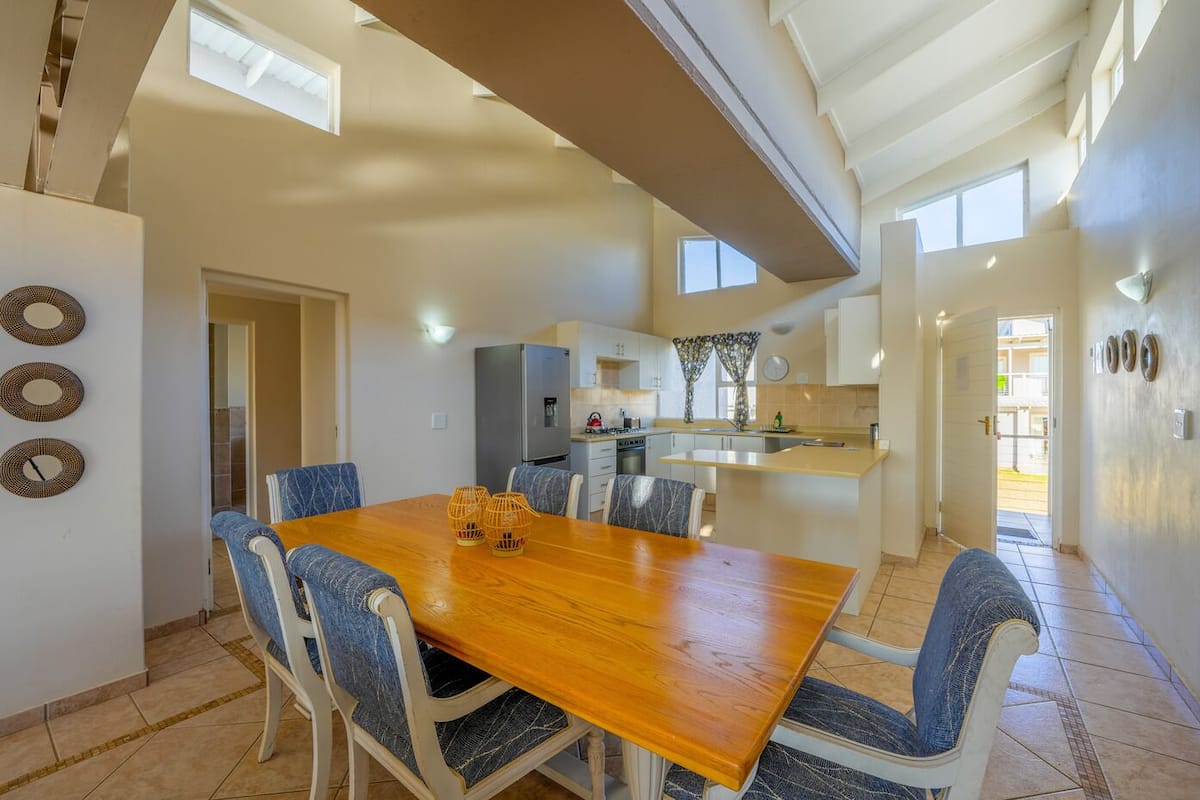Uma sala de jantar espaçosa com uma mesa de madeira e cadeiras estofadas em azul, uma cozinha com geladeira e pia, e vista para o exterior por uma porta.