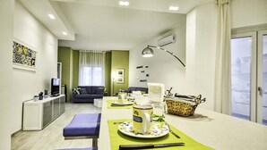 A modern living room with a sofa, a dining table set with a teapot, cups, and a knife. There is a small kitchen area with a refrigerator and a TV. - Casa Primavera with shared pool - My Rental Homes (Sorrento)