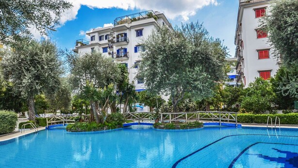 A swimming pool with a wooden deck and surrounding greenery, set against a backdrop of multi-story buildings with balconies. - Casa Primavera with shared pool - My Rental Homes (Sorrento)