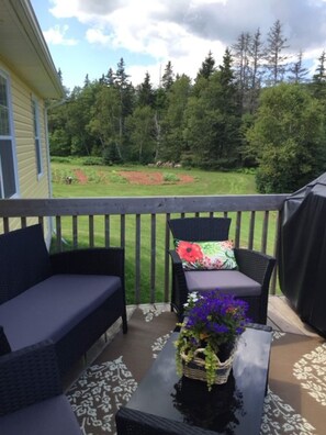 A covered outdoor seating area with two wicker sofas and a table, overlooking a landscaped garden and forested area.