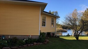 A two-story house with a yellow siding exterior, a front porch with a small garden, and a view of a lake. - Beautiful water's edge 2 bedroom vacation home (Breadalbane)
