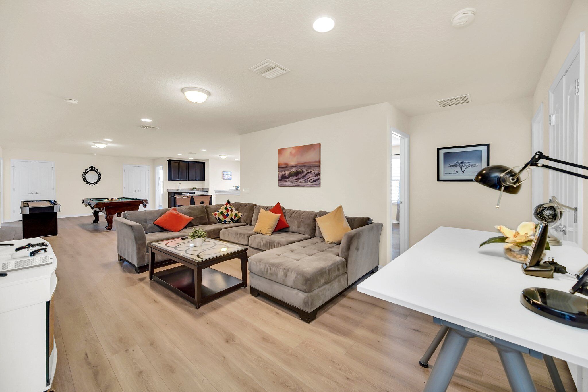 A spacious living room with a sectional sofa, a coffee table, and a dining table. There is a pool table in the background, and the room has a modern design with white walls and wooden flooring.