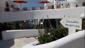 Ein Empfangsbereich mit einem Schild, das 'RECEPTION' in Englisch und einer weiteren Sprache anzeigt. Im Hintergrund sind zwei rote Sonnenschirme und ein Pool zu sehen.