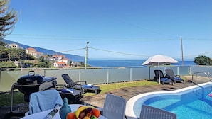 Ett område vid poolen med utsikt över havet, en grill, solstolar och ett bord med frukt och en drink.