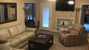 Un salon avec un canapé beige, un fauteuil à motifs et une table basse. Un téléviseur à écran plat est fixé au mur, et un ventilateur de plafond est visible.