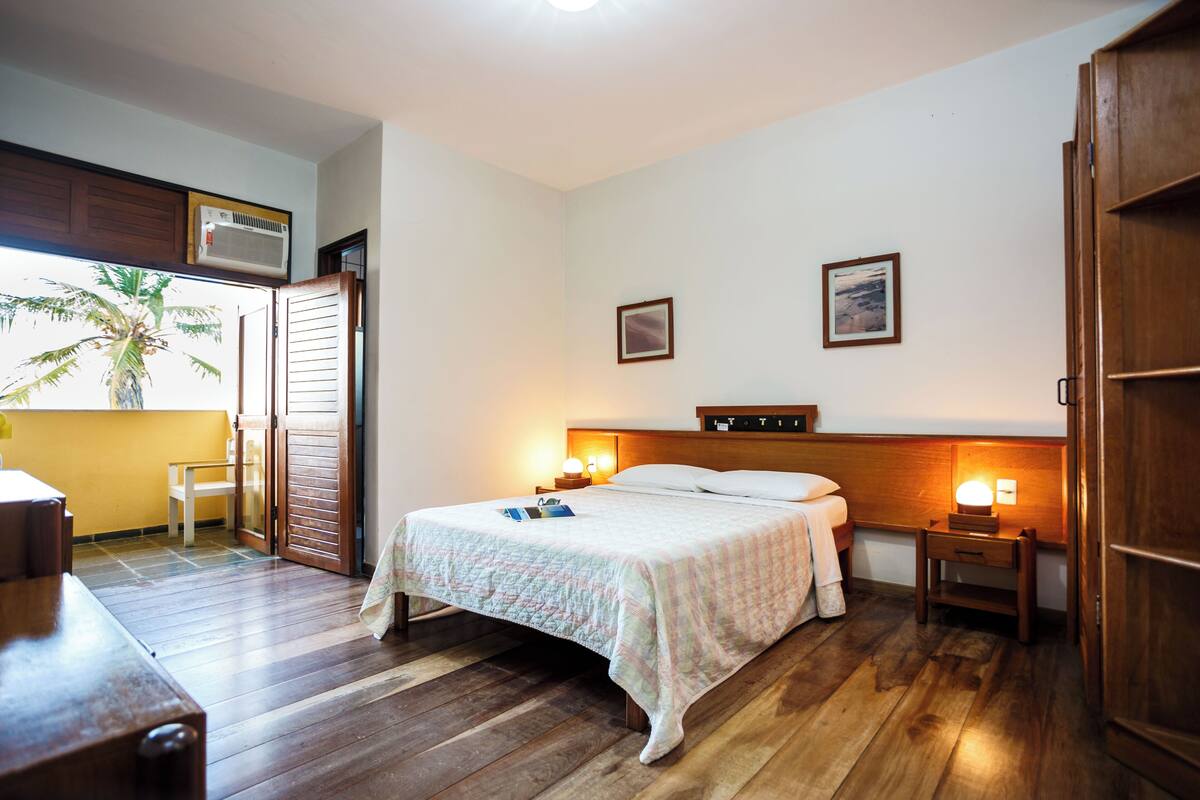 Un dormitorio con cama de madera, lámparas de noche, mesita de noche, armario de madera y ventana con vista a palmeras.