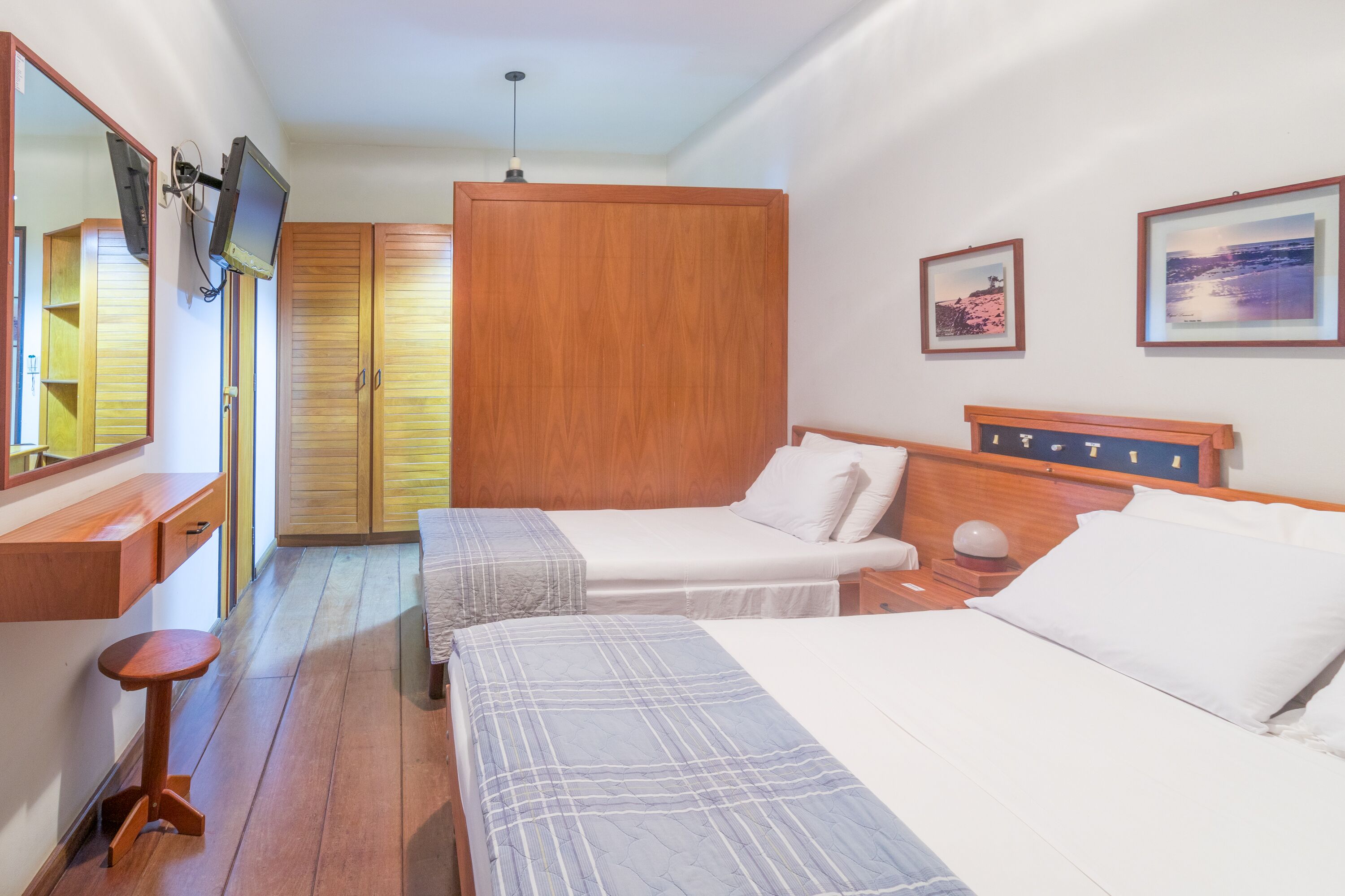 A hotel room with two beds, wooden wardrobe, a TV mounted on the wall, and framed pictures on the wall.