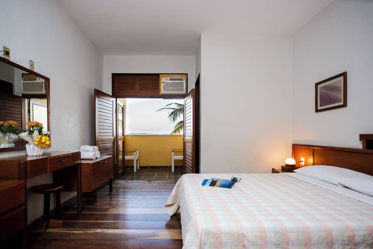 Habitación de hotel con cama, mesita de noche y escritorio con silla. Se ve una ventana con vista a palmeras y un balcón.