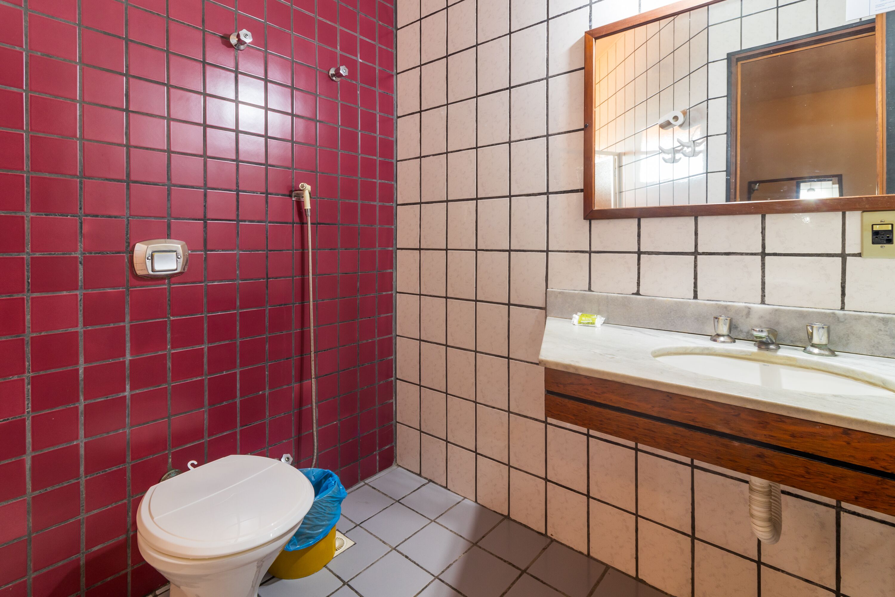 Un baño con pared de baldosas rojas, inodoro blanco, lavamanos con espejo y zona de ducha.