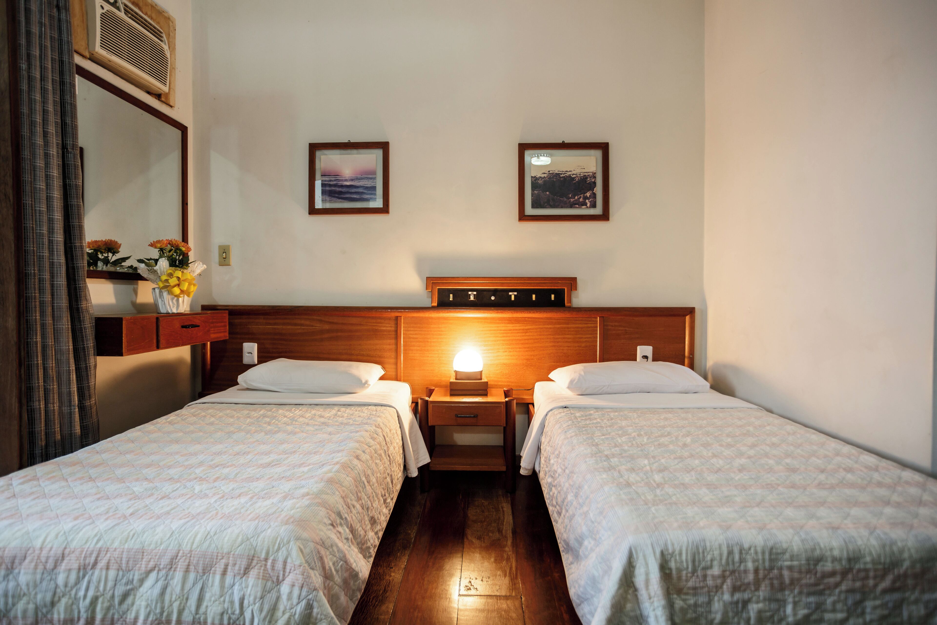 A hotel room with two single beds, a nightstand with a lamp, a mirror, and two framed pictures on the wall.