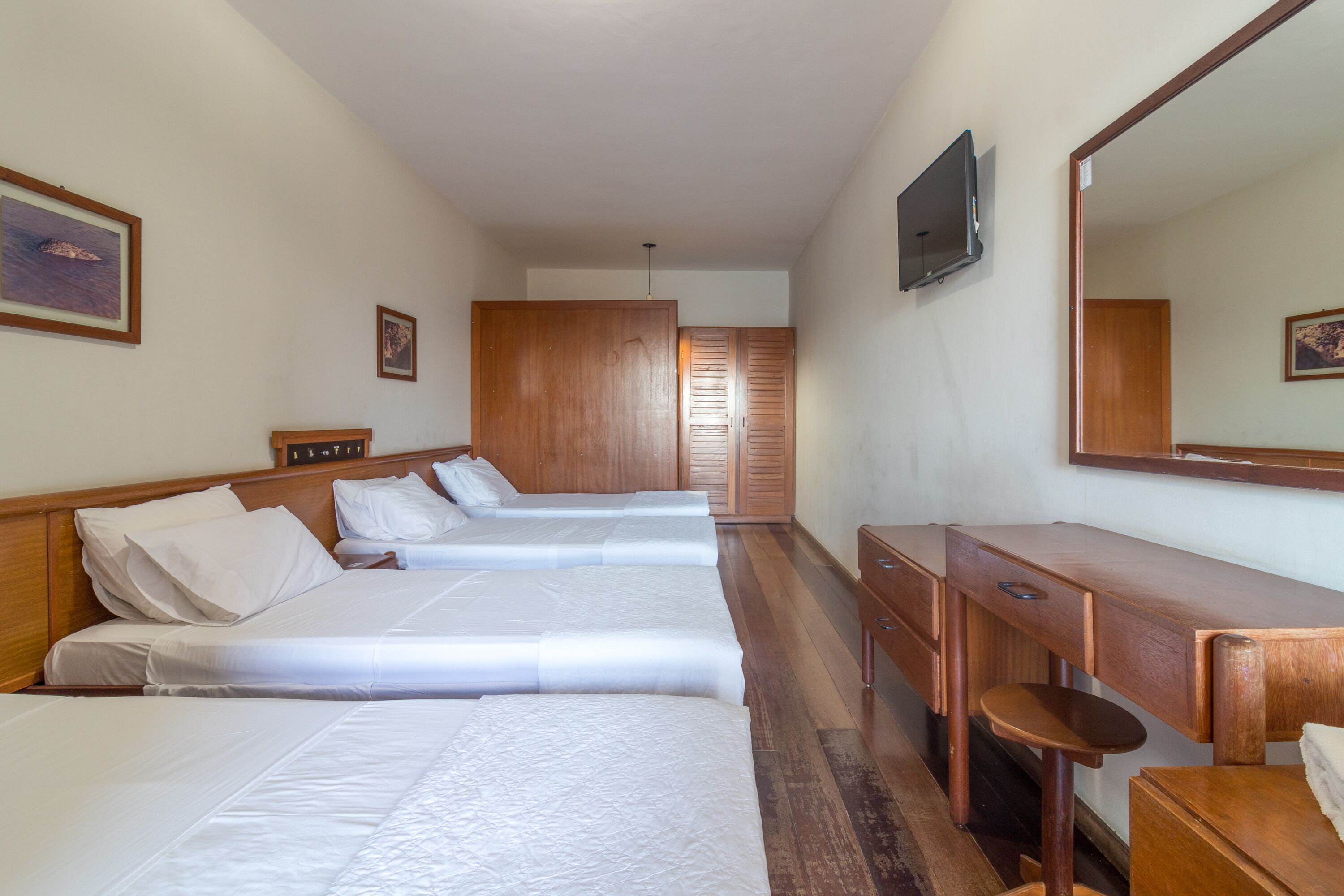 Habitación de hotel con tres camas, un escritorio de madera, una silla, un televisor y un armario.