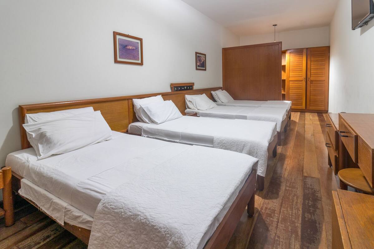 Una habitación de hotel con tres camas individuales, suelo de madera, un armario de madera y una mesita de madera.