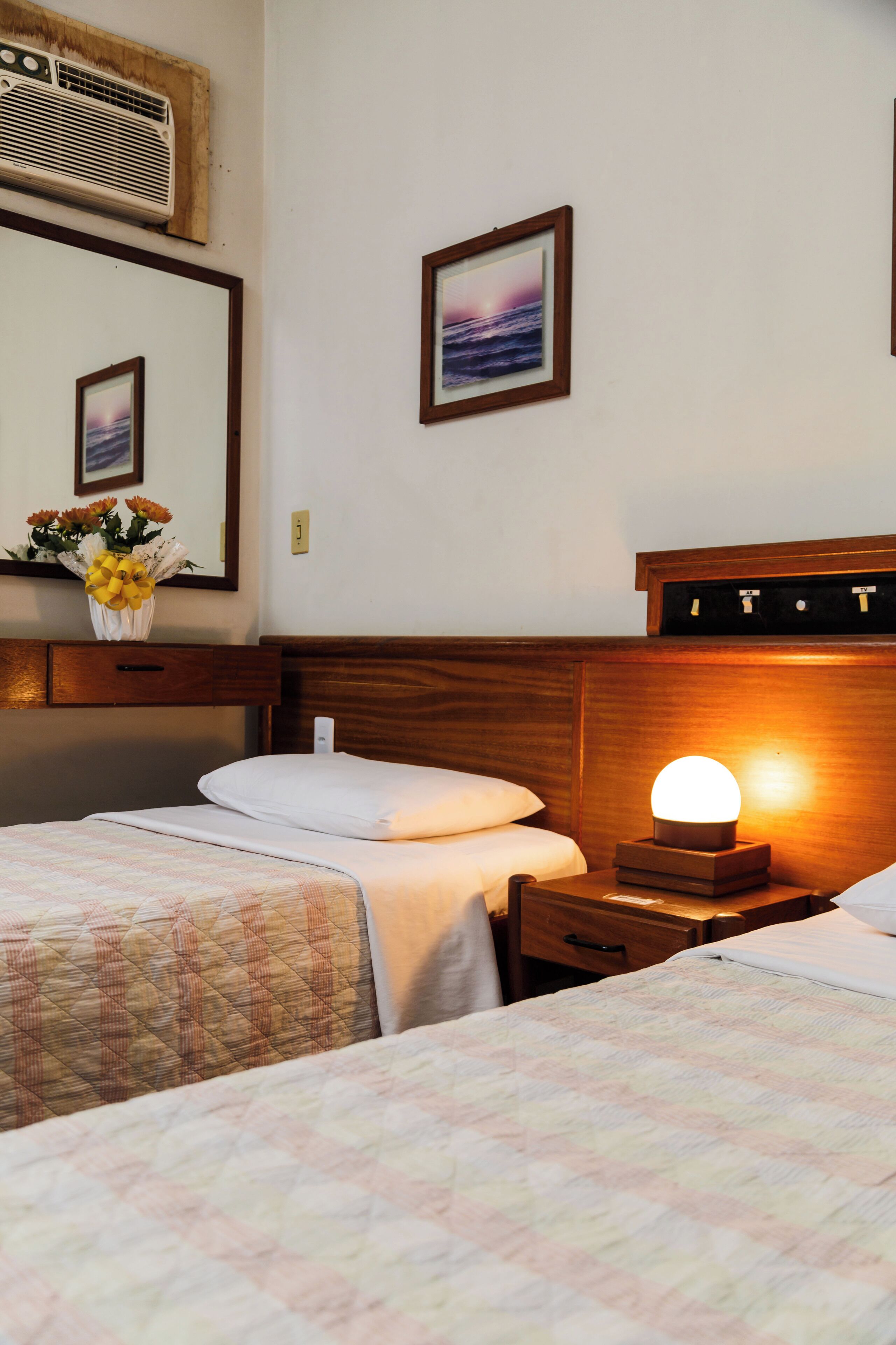 Habitación de hotel con dos camas, una mesita de noche con lámpara, un espejo y un aire acondicionado instalado en la pared.