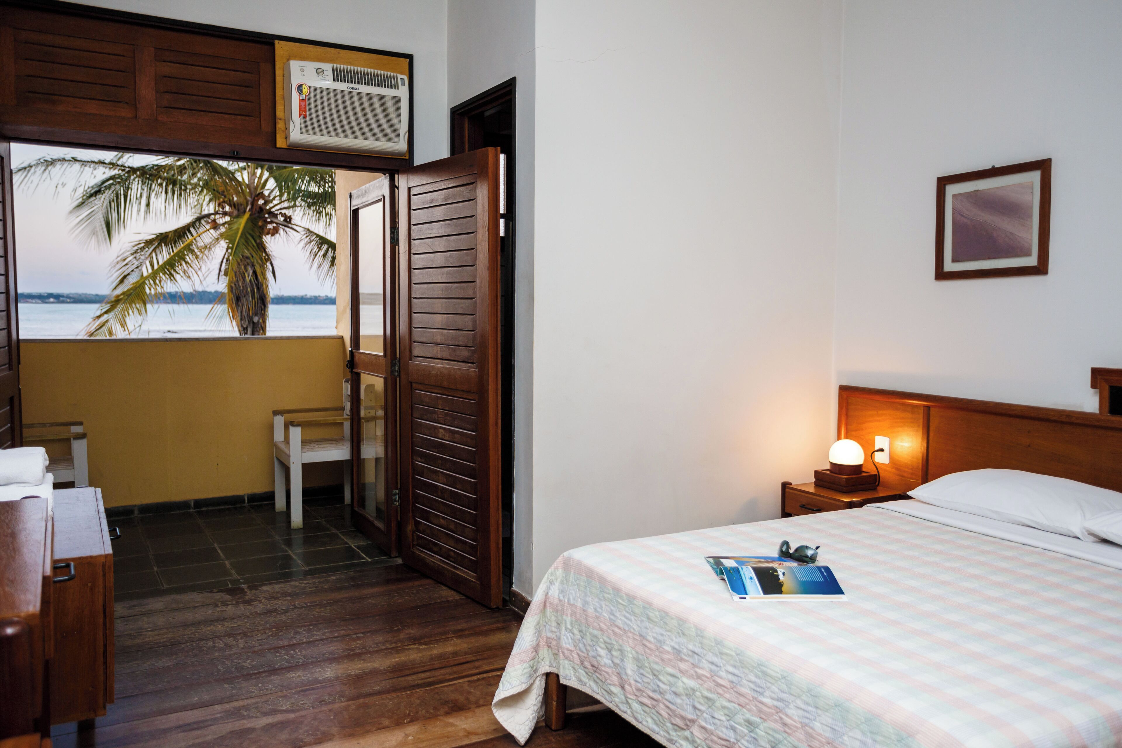 A hotel room with a bed, a nightstand, a lamp, a wooden door, a chair, and a view of the ocean and palm tree.