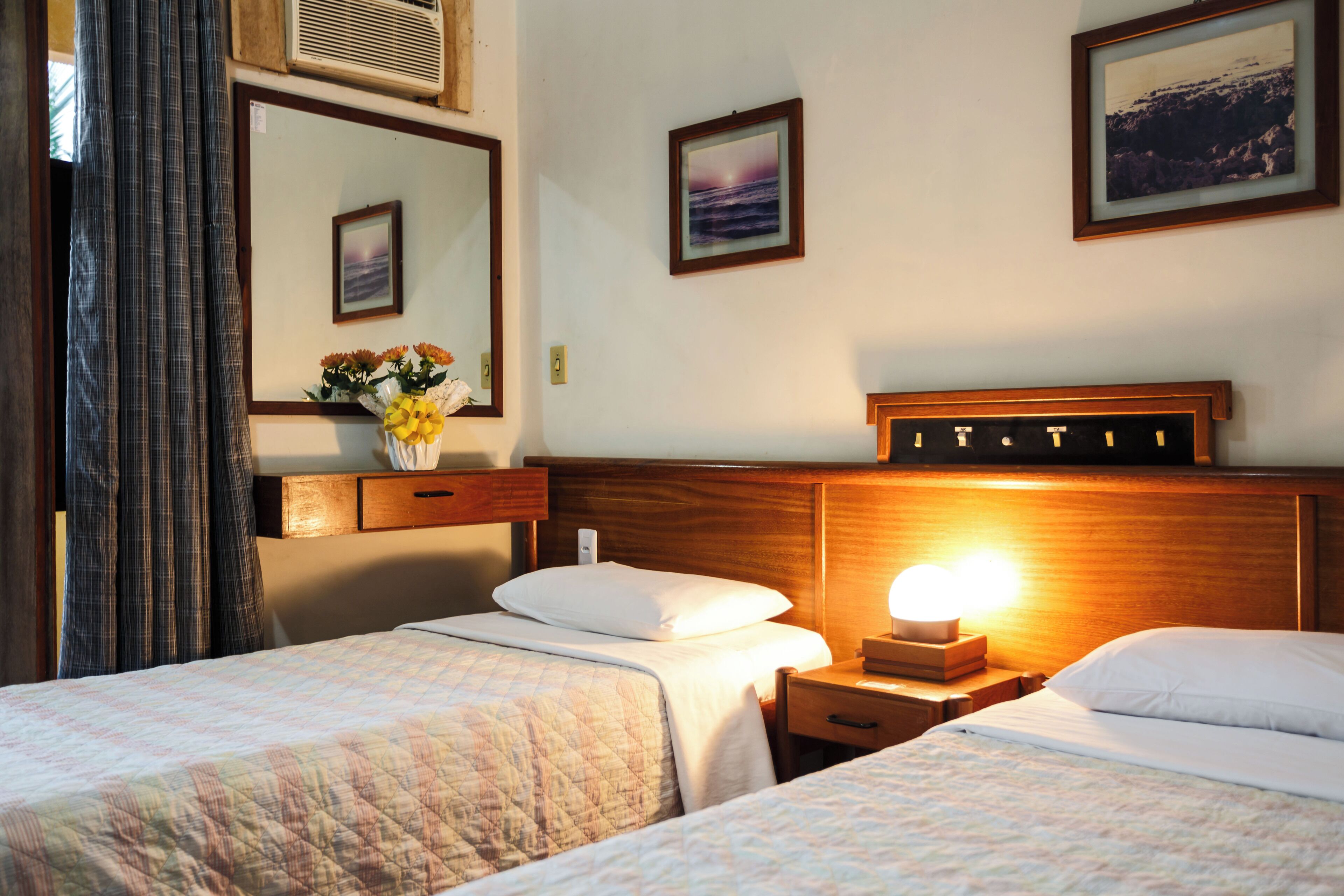 A hotel room with two beds, a nightstand with a lamp, a mirror, and framed pictures on the wall.