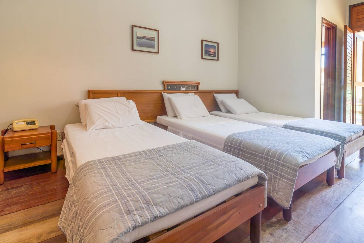 Una habitación con tres camas individuales, cabeceros de madera, una mesita de noche con lámpara y dos cuadros enmarcados en la pared.