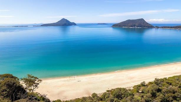 A coastal landscape with a sandy beach, clear turquoise waters, and two prominent hills in the distance. - Columbia Views,  2/9 Columbia Close - Aircon, WIFI, Netflix, Views & Boat Parking (Nelson Bay)