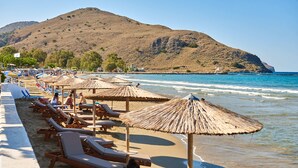 A beach with rows of sun loungers and thatched umbrellas, a sandy shore, and a mountain in the background. - Lake View Suites (Apokoronas)