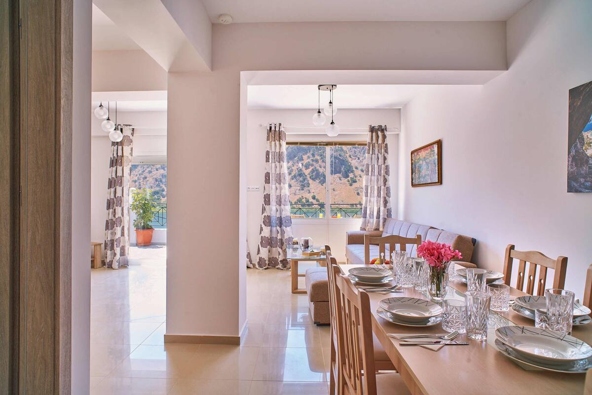 A dining area with a long table set for a meal, a sofa, and a view of a mountainous landscape through the window.