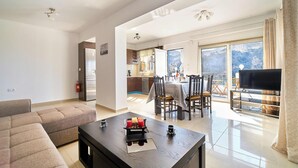A spacious living area with a sofa, a coffee table, and a TV. There is a balcony with a view of the mountains. - Lake View Suites (Apokoronas)