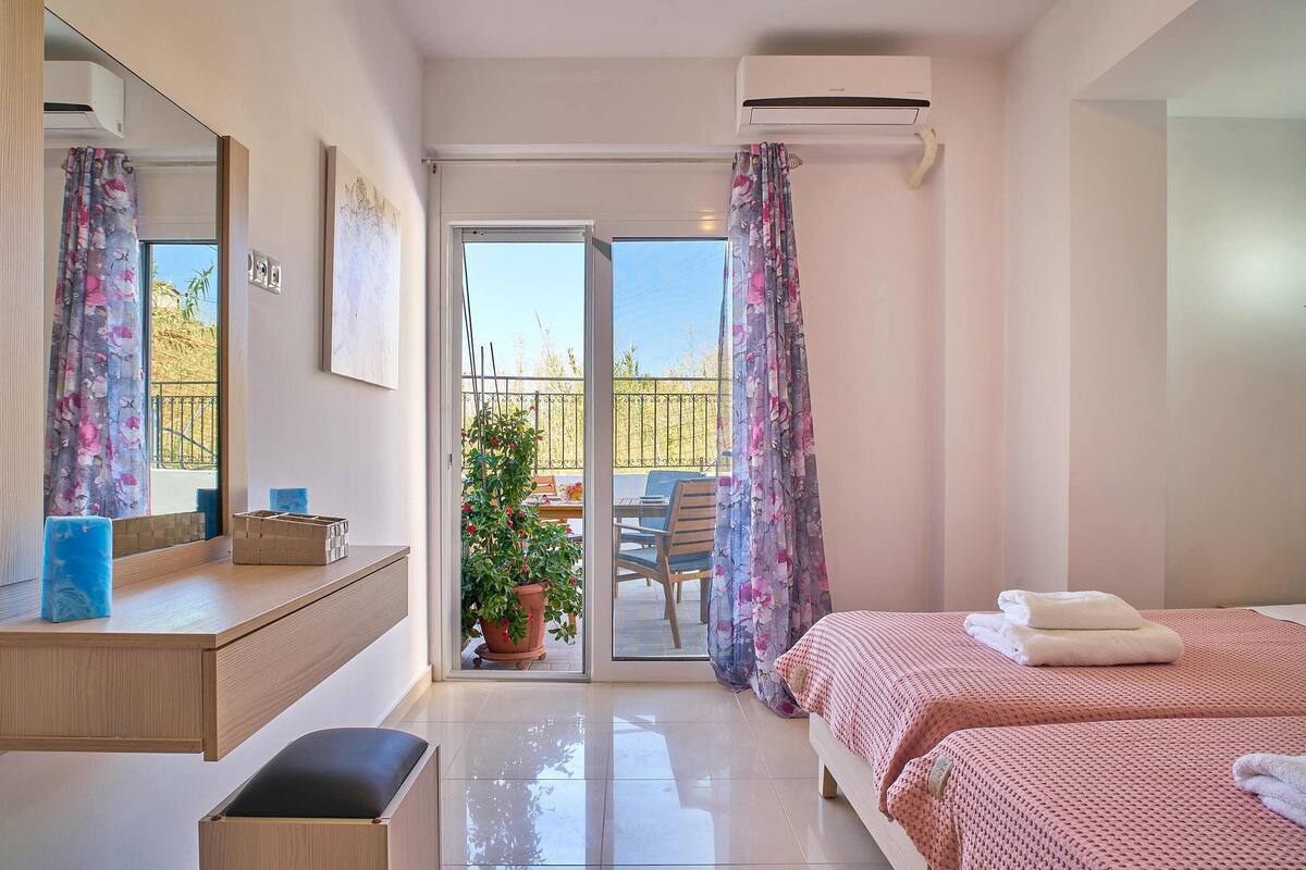 A hotel room with a sliding glass door to a balcony, a wooden desk, a bed with pink bedding, and a potted plant.