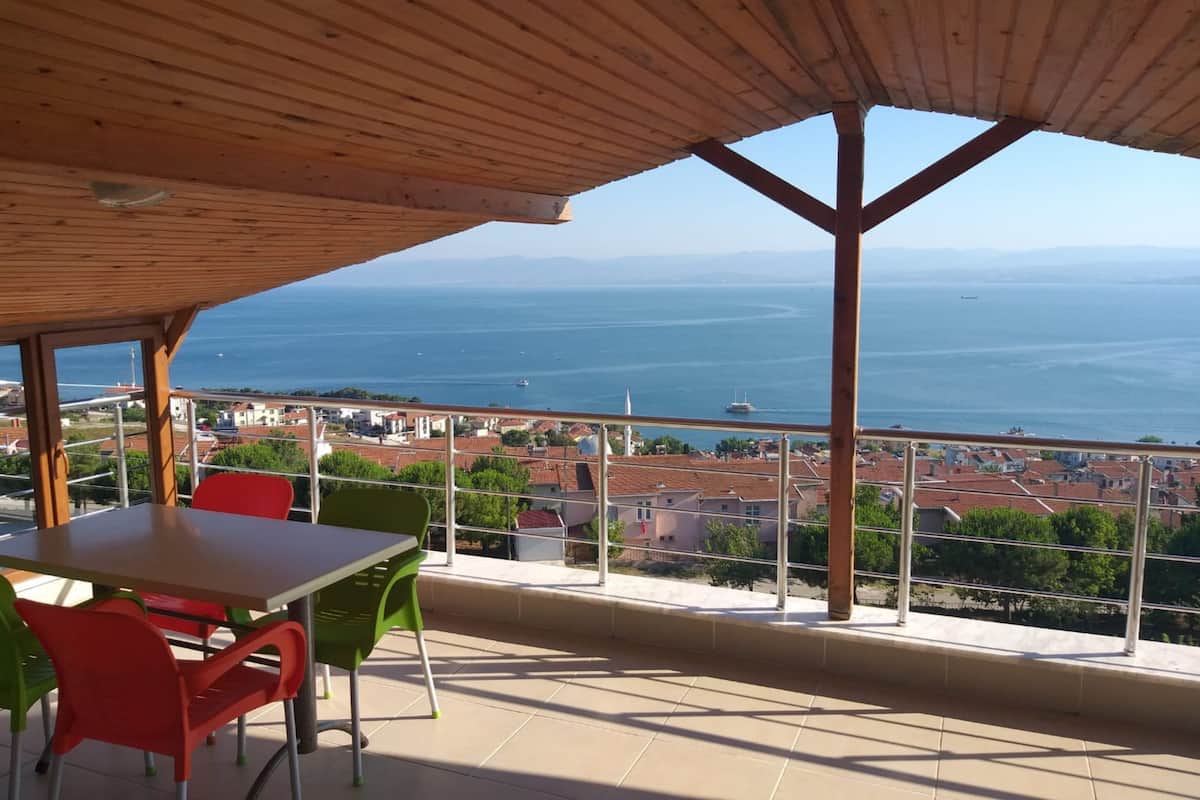 A terrace with a wooden roof, a table, and chairs. It overlooks a coastal town with buildings and a view of the sea.