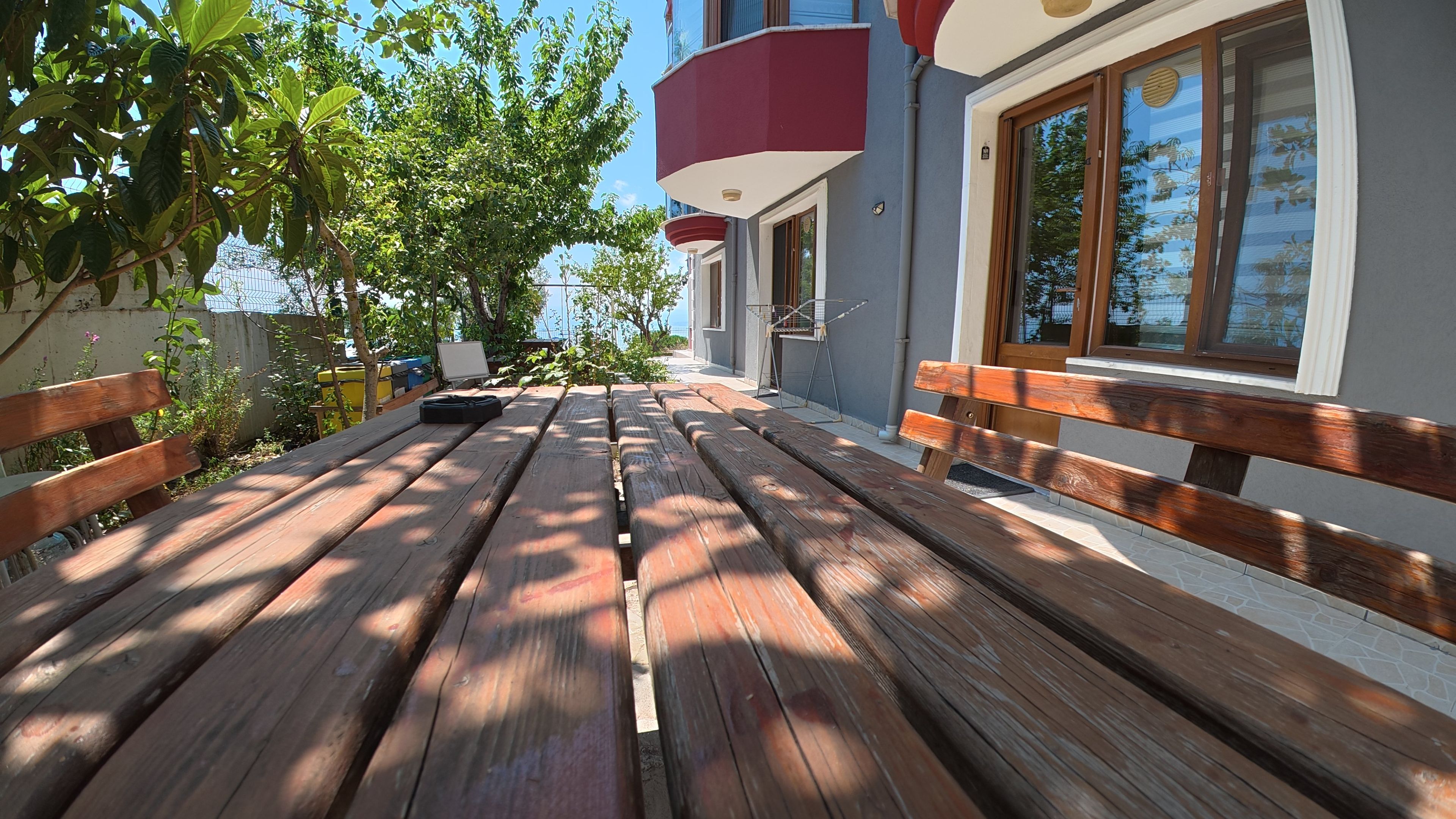 Terraza de madera al aire libre con bancos, rodeada de follaje y un edificio con un balcón rojo.