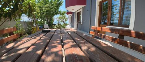 Terraza de madera al aire libre con bancos, rodeada de follaje y un edificio con un balcón rojo.